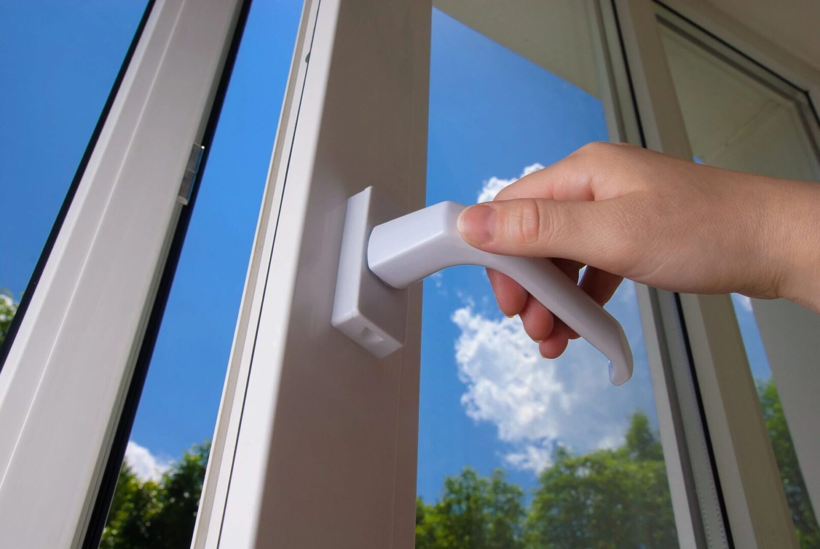 A person's hand opening a white window handle with a view of a blue sky and green trees outside.