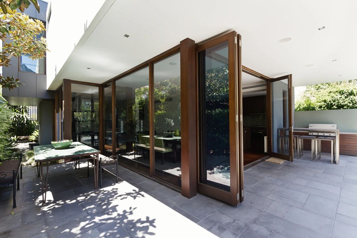 Modern patio with large folding glass doors, wooden frames, and a tiled floor, leading into a sunny, landscaped garden.