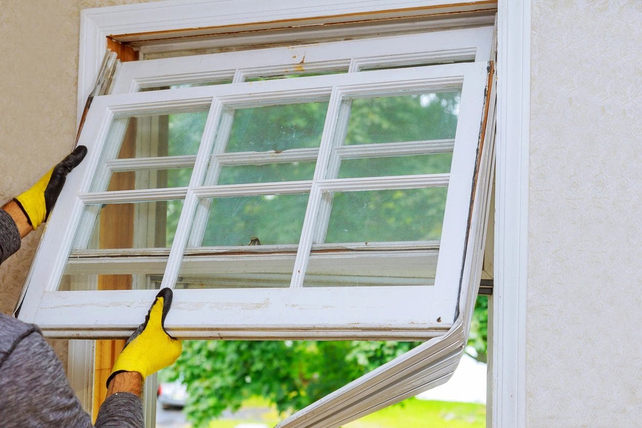 Person wearing gloves installs a new white window frame into a wall, enhancing the building's exterior aesthetics.