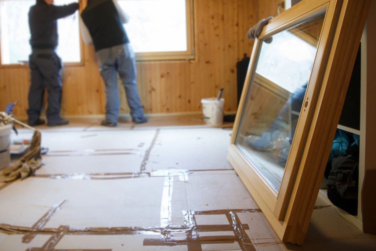 Two people installing a window in a room covered with protective sheets, focusing on work near a wooden wall.