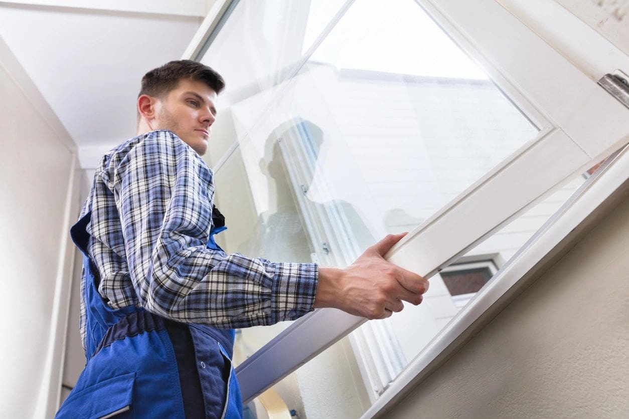 A man in a plaid shirt and blue overalls installing a window in a bright, modern room.